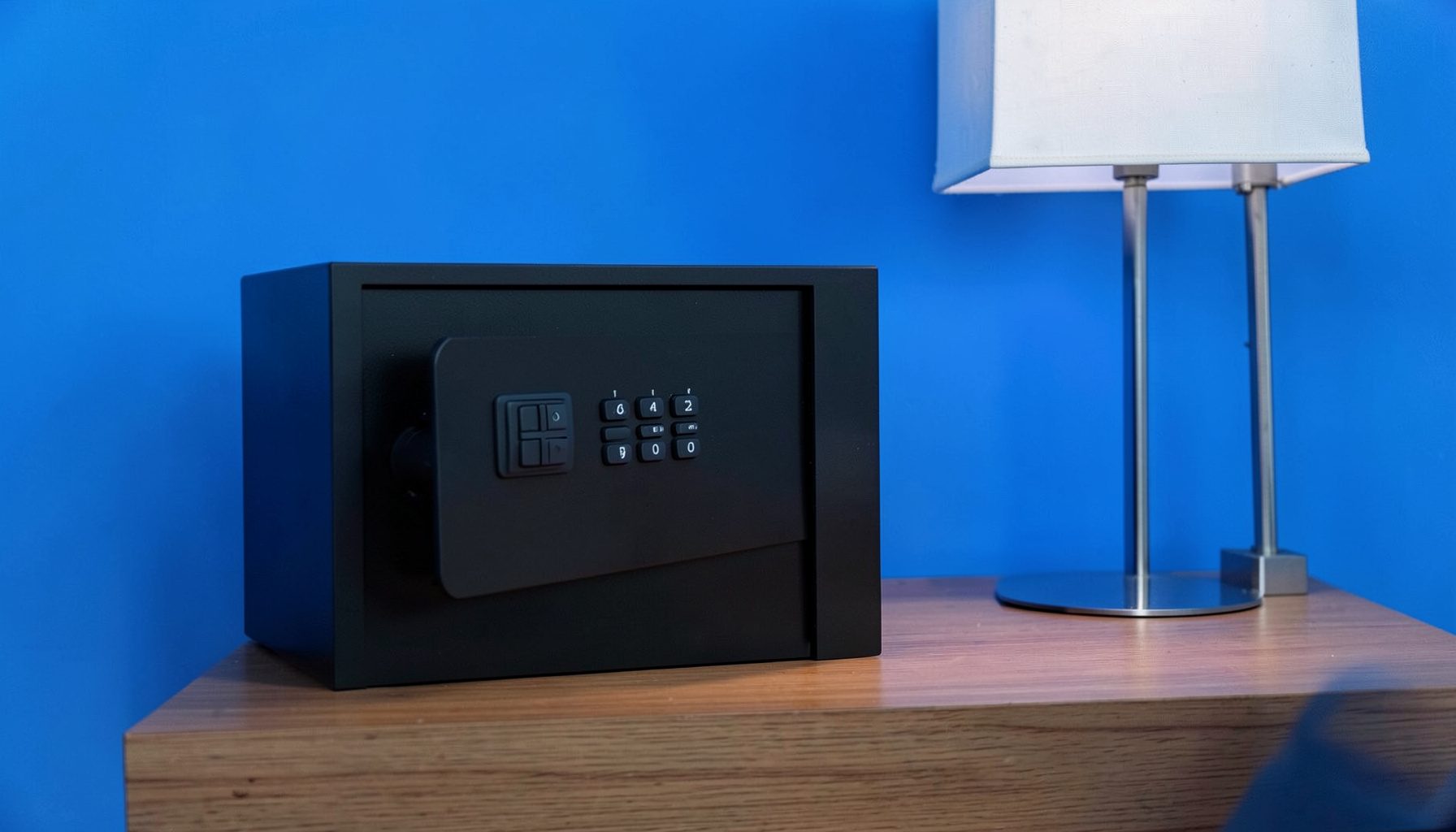 A low-profile bedside gun safe in matte black with a digital keypad, placed on a wooden nightstand beside a lamp