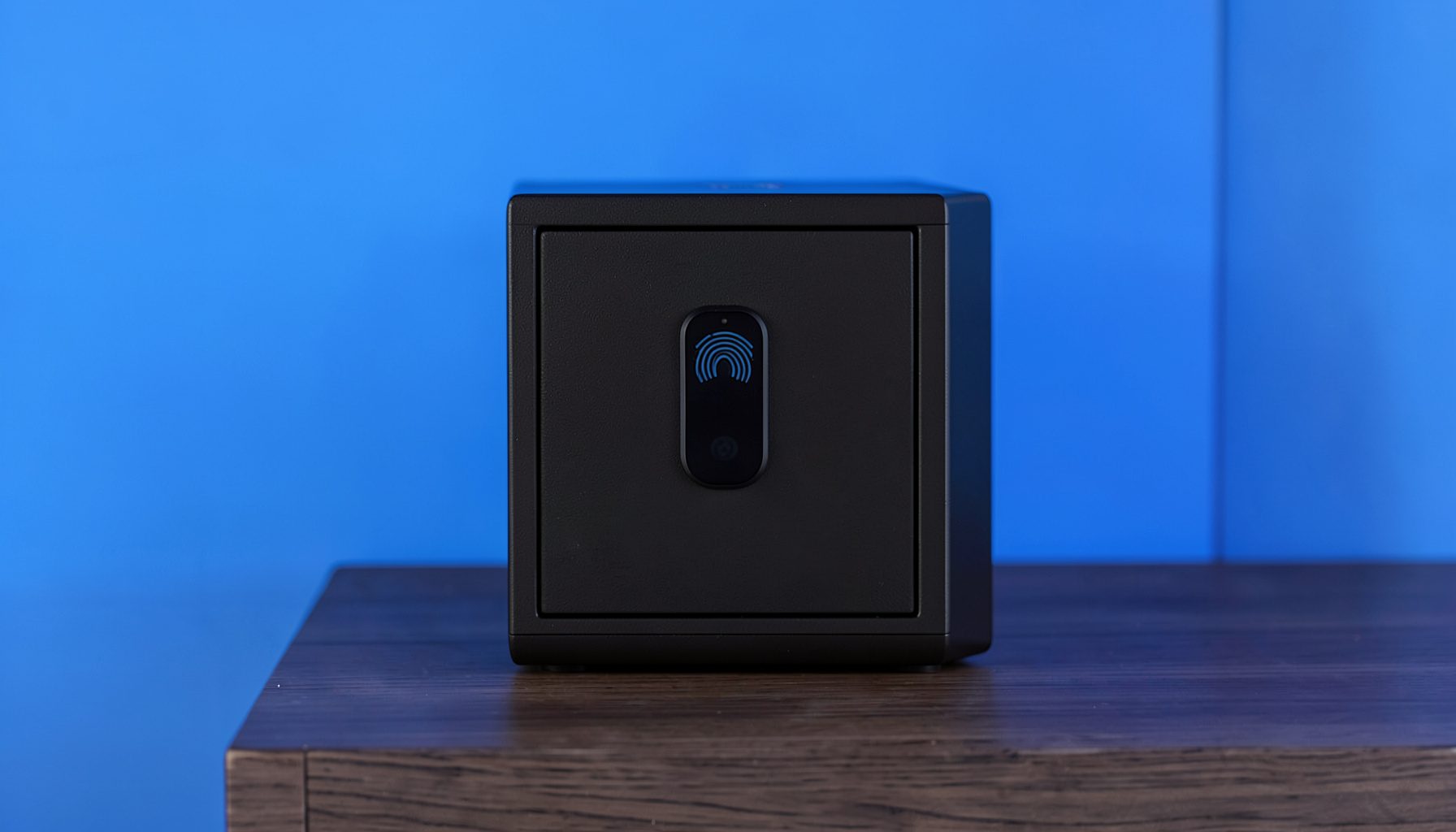 A compact biometric gun safe with a sleek fingerprint scanner on the top panel, dark matte black finish, sitting on a nightstand