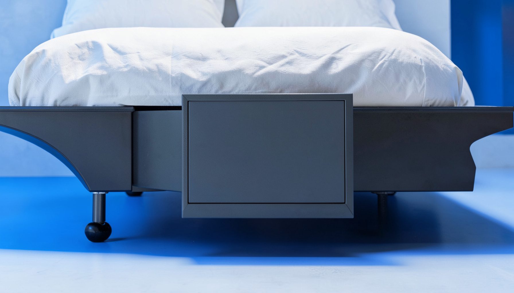 A low-profile underbed gun safe on casters, dark gray steel, partially pulled out from beneath a bed frame