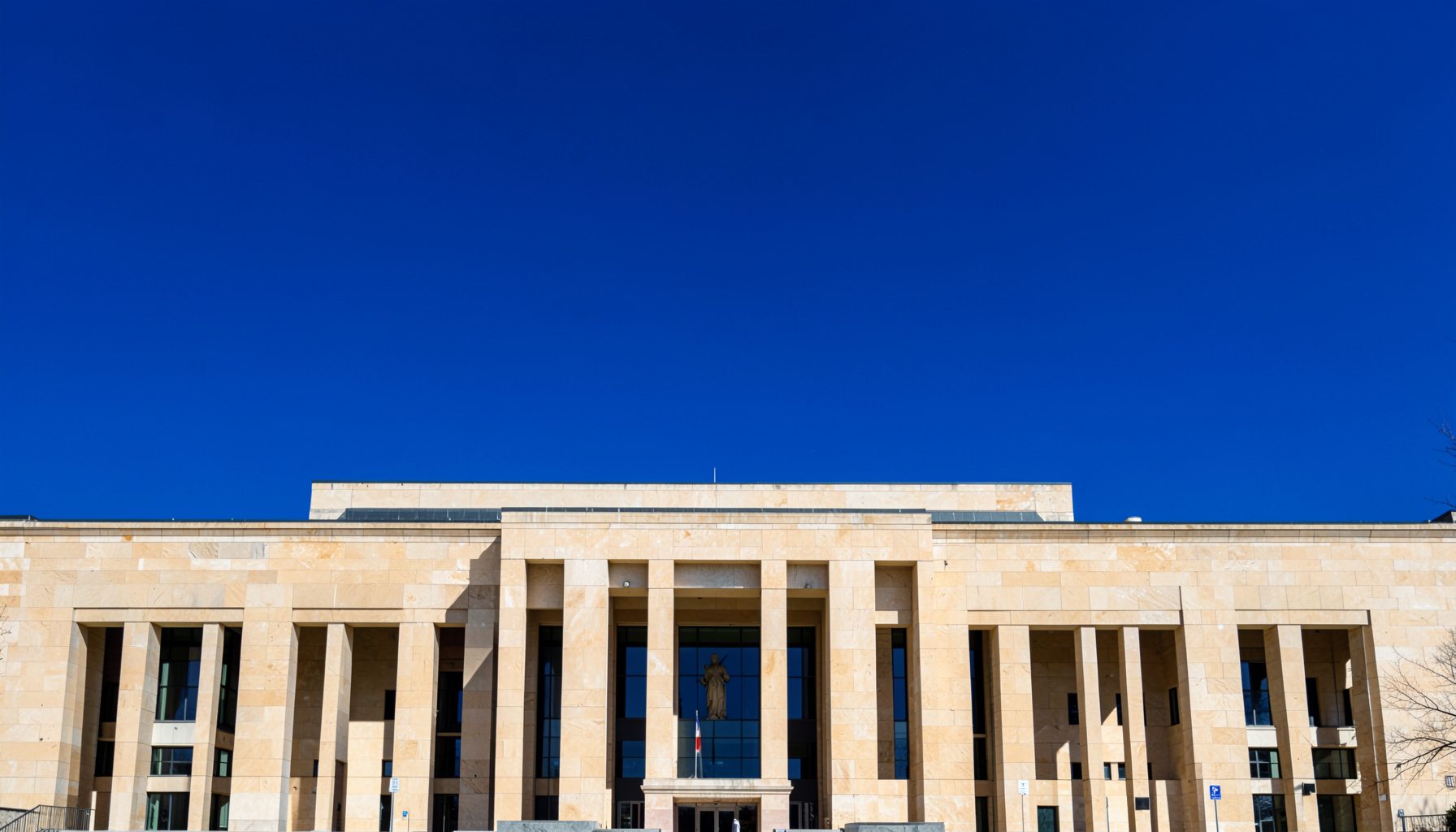 A clean architectural photograph with strong symmetry: a wide, low-profile government building facade in warm beige stone, framed by a deep blue sky, conveying order and civic authority.