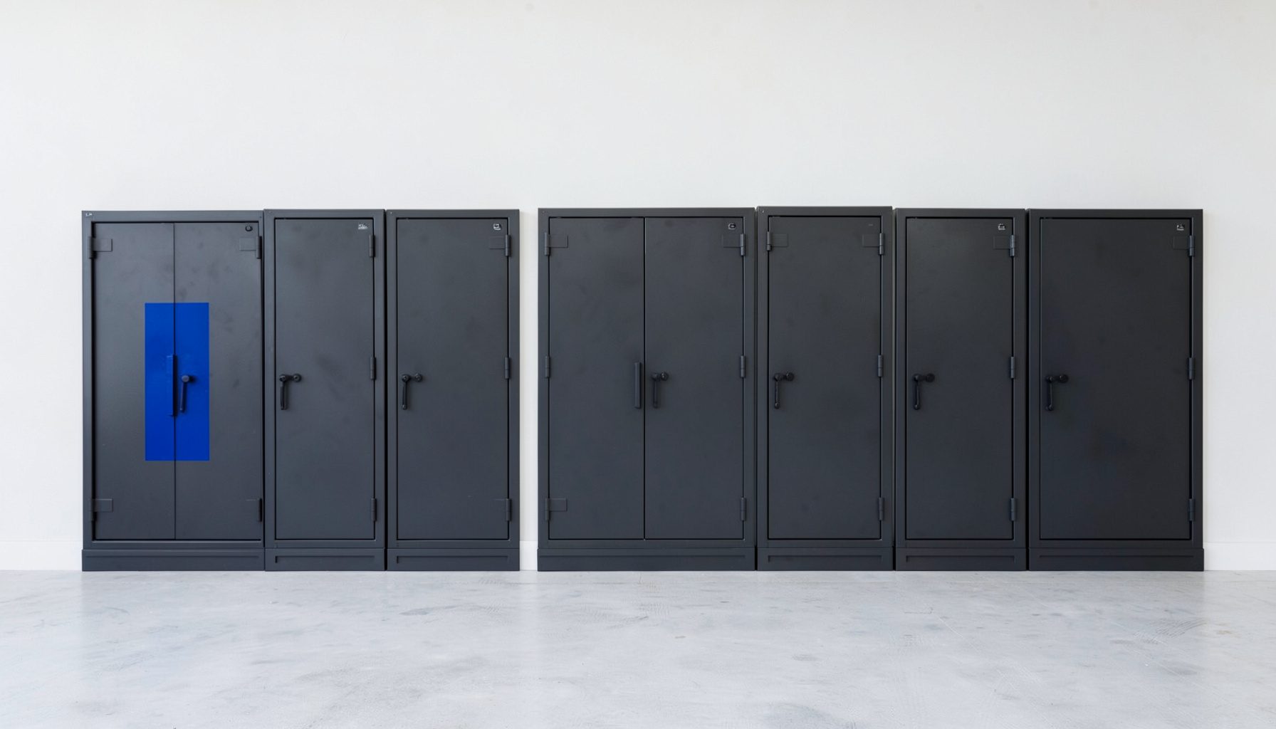 A clean architectural photograph of a row of dark steel gun safes lined against a pale wall, evenly spaced with strong symmetry, calm and orderly atmosphere.